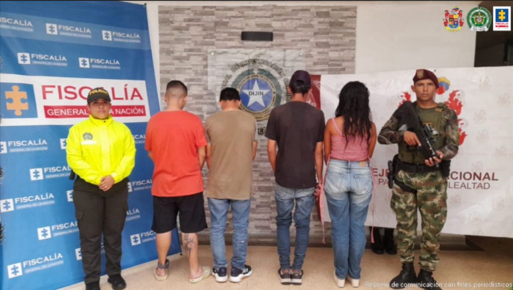 En la imagen se observa a cuatro capturados de espaldas junto a dos uniformados de la Policía y Ejército Nacional. Detrás de ellos los banners que identifican a la Fiscalía General de la Nación y Ejército Nacional.