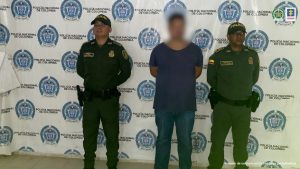 En la imagen se observa al investigado de frente con el rostro difuminado junto a dos uniformados de la Policía Nacional. Detrás de ellos el banner que identifica a la Policía Nacional.