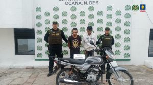Alternativo: Se visualiza a los capturados junto a dos uniformados de la Policía Nacional. Frente a ellos la motocicleta que se recuperó tras su hurto. Detrás el banner que identifica a la Policía Nacional en Ocaña