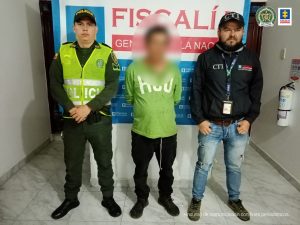 en la imagen se ve una persona detenida bajo cutodia de un investigador del CTI y de la Policía Nacional. detrás suyo un backing de la Fiscalía.