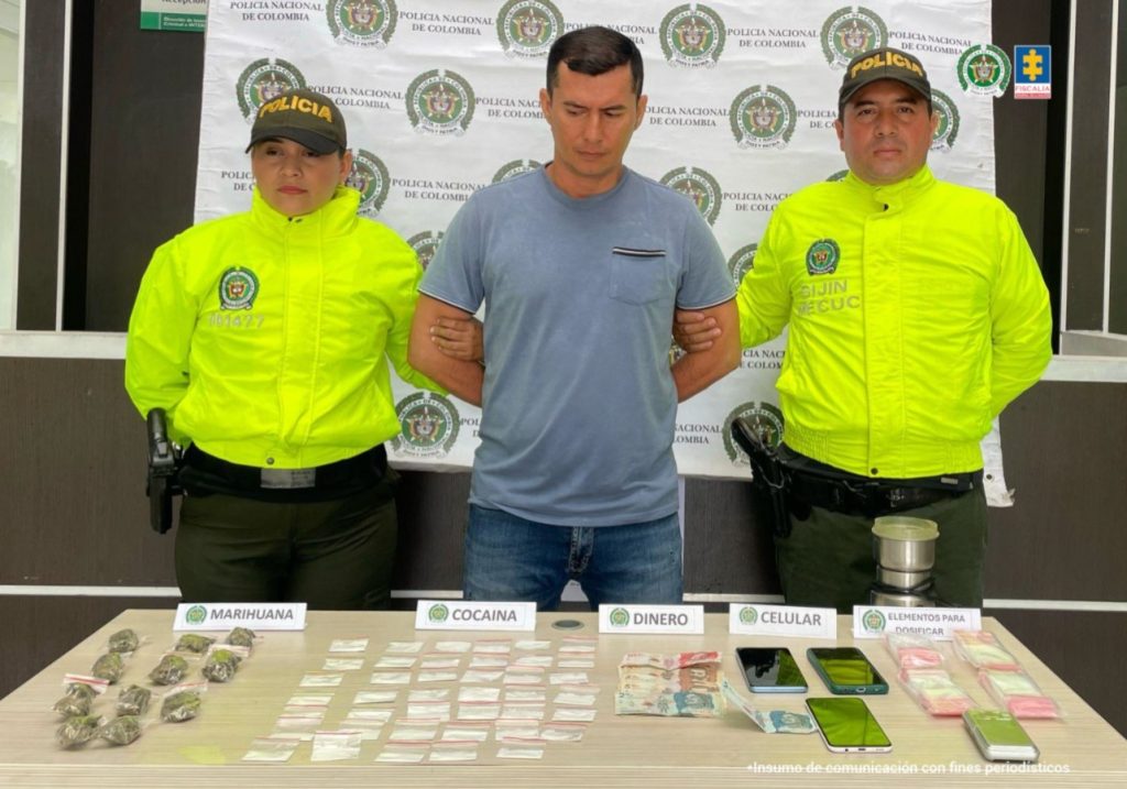 Se visualiza al capturado junto a dos uniformados de la Policía Nacional. Frente a ellos una mesa con el material incautado. Detrás el banner que identifica a la Policía Nacional.